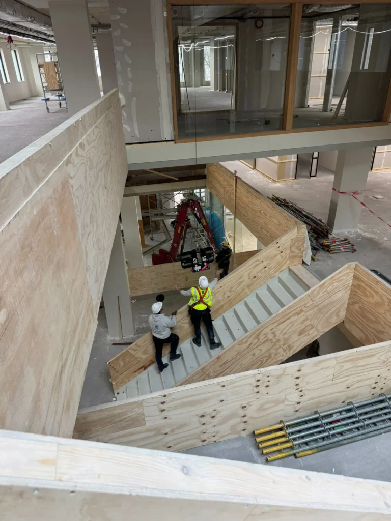 Twee werklui op houten trap begeleiden glasplaatsing met glaszuiger in trappengat